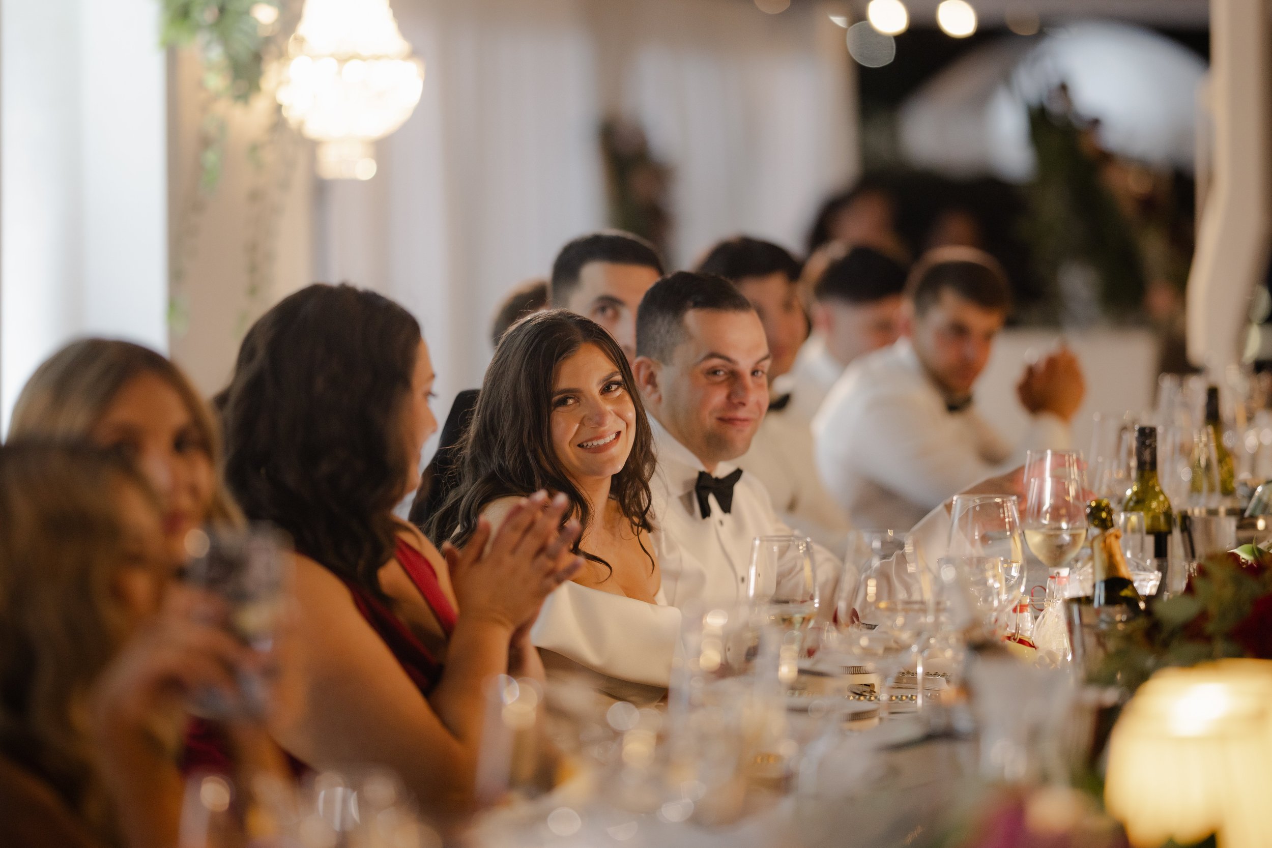 A radiant bride smiles warmly during reception speeches at a candlelit wedding table, captured in cinematic editorial style.