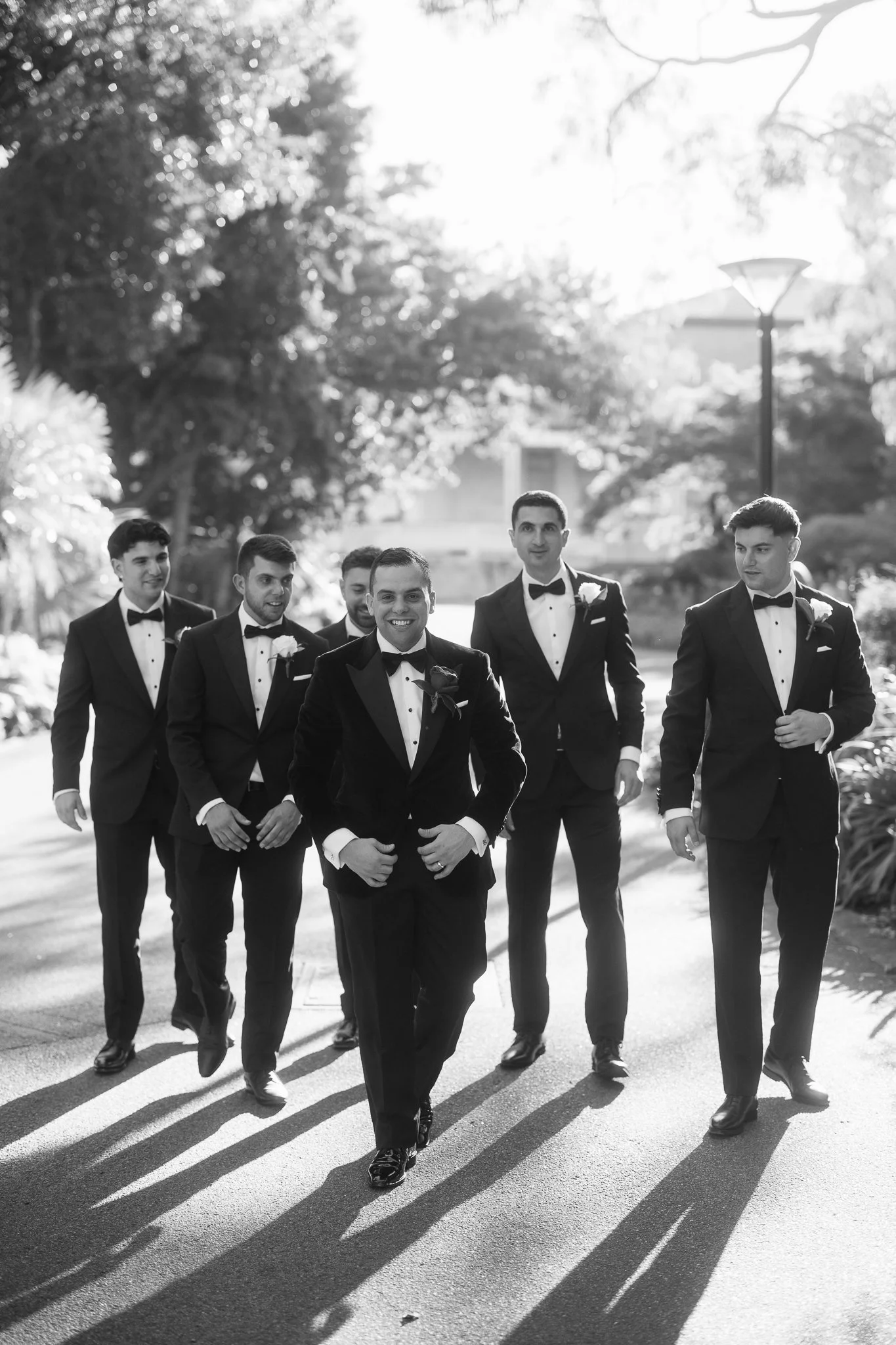 Smiling groom leads his groomsmen in matching black tuxedos along a sunlit tree-lined path on his wedding day.