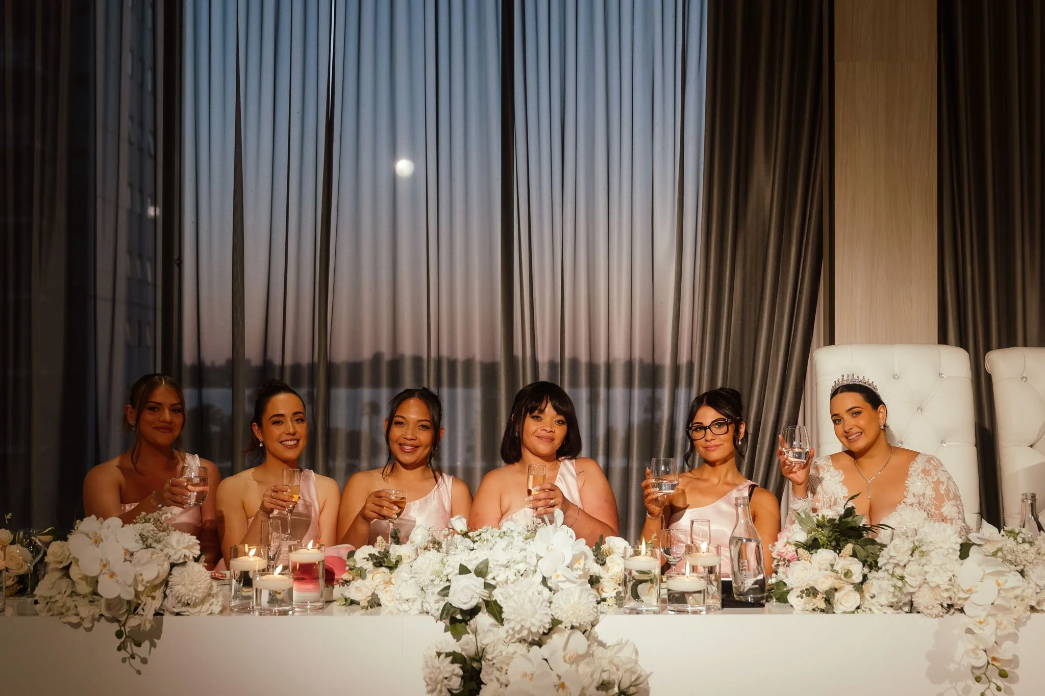 Bride wearing a crystal tiara and five bridesmaids in blush pink gowns raising glasses in a toast at the Crown Perth bridal table, with a full moon visible through the floor-to-ceiling windows behind them, photographed by James Di Stefano