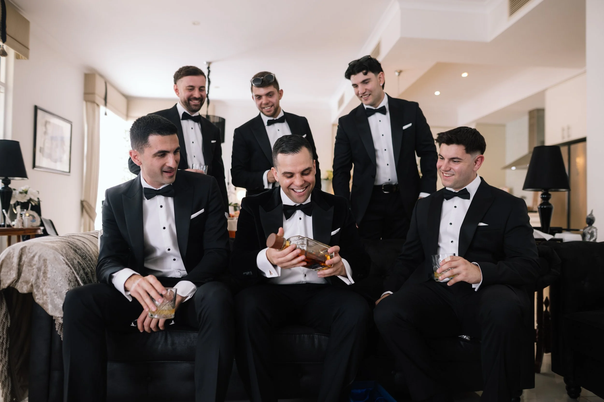 Groom laughing with his groomsmen in black bow-tie tuxedos sharing a whisky toast during wedding morning preparations in Perth