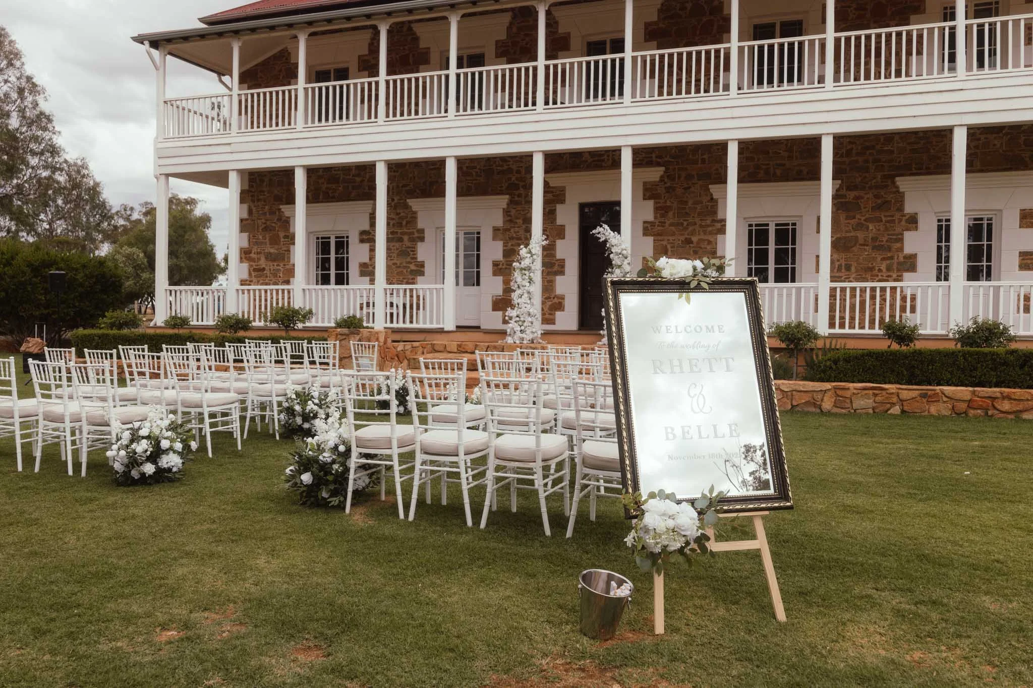 A wide photograph capturing the outdoor ceremony lawn at Buckland Estate, a hidden outback wedding venue near Perth. White chairs are neatly arranged facing a large mirror sign, all positioned in front of the grand heritage homestead.