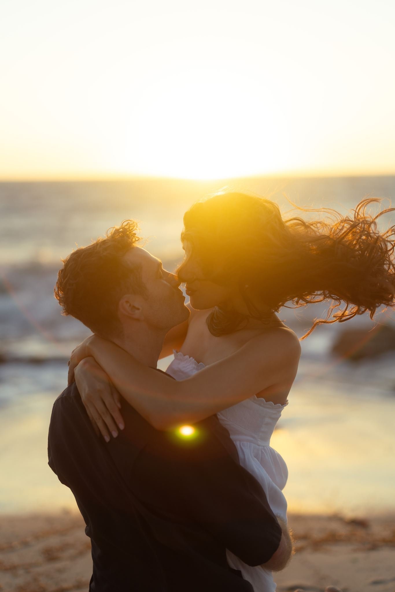 A groom lifts his bride-to-be for a windswept kiss against a blazing golden-hour sunset on a Perth beach during their pre-wedding shoot