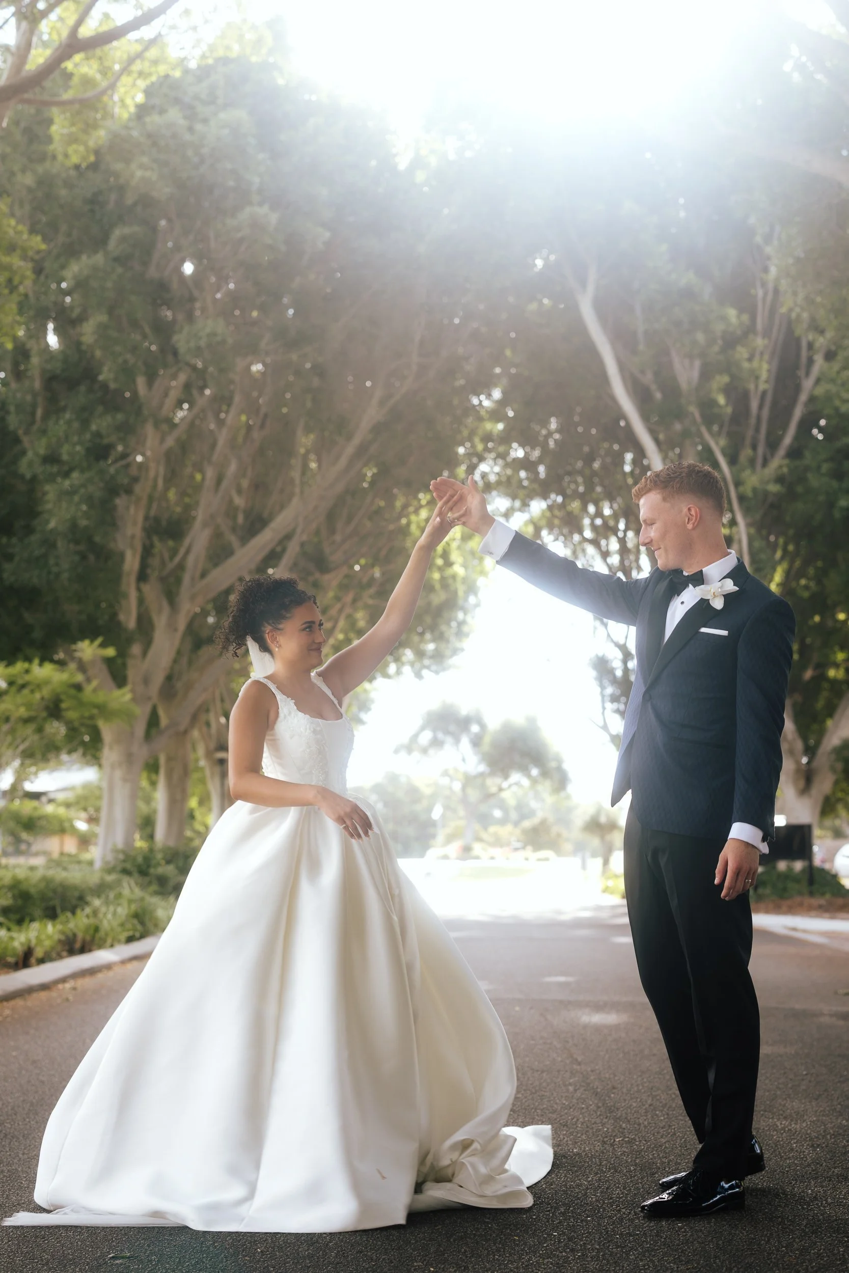 Groom spinning bride in a full satin ball gown along a sun-drenched tree-lined driveway during their wedding portraits.