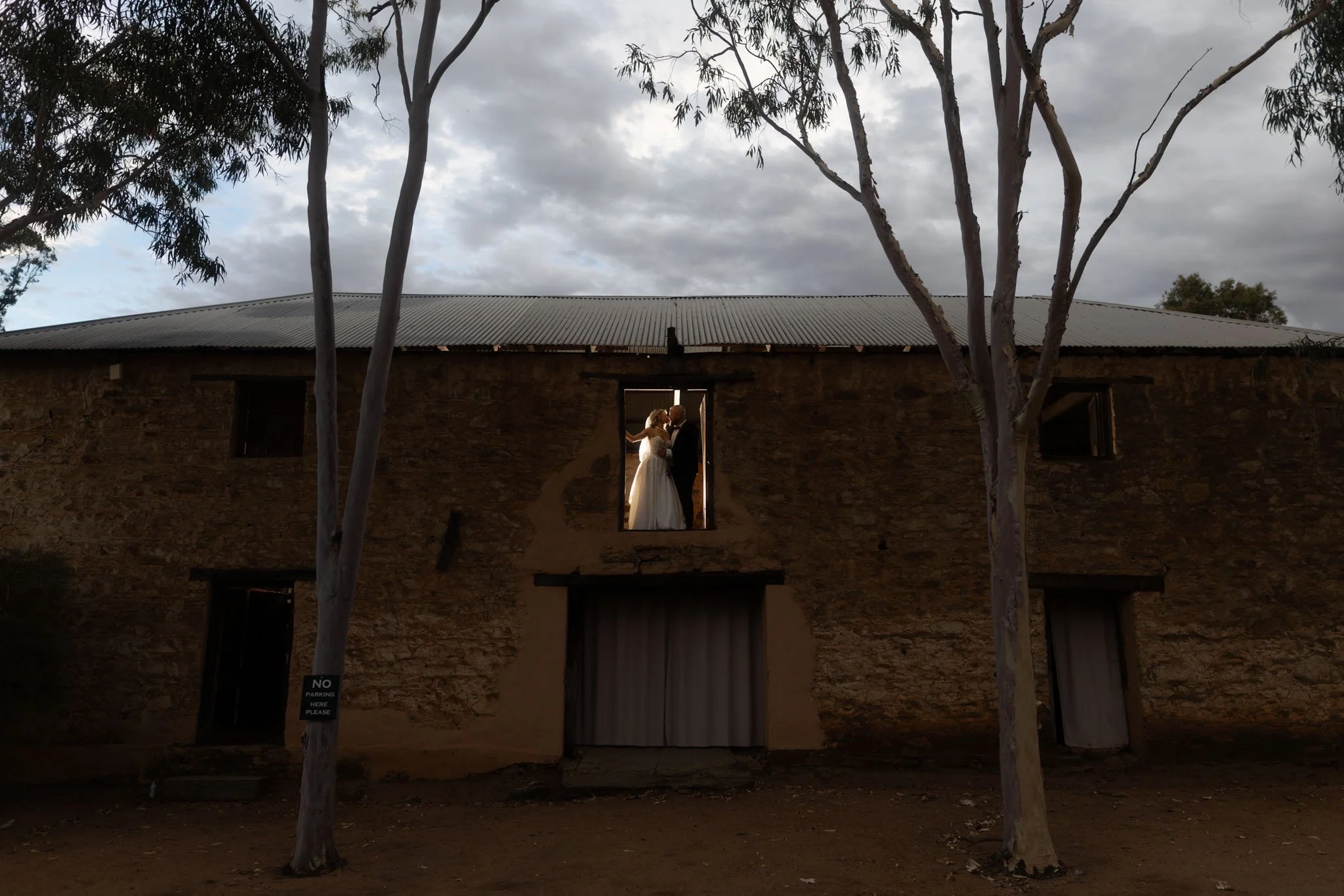 A dramatic, wide architectural wedding photo of Buckland Estate's historic stone granary. The bride and groom are beautifully backlit as they embrace in an elevated loft doorway, framed by towering outback trees and an moody sky