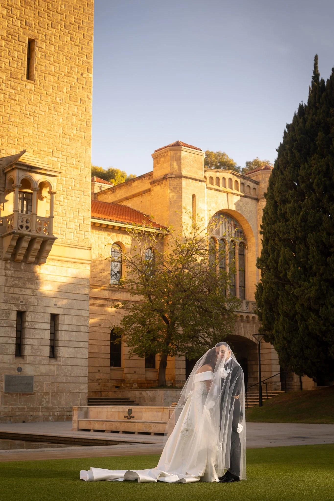 Bride and groom share a kiss beneath a sweeping cathedral veil on the lawn of a golden sandstone heritage building at sunset.