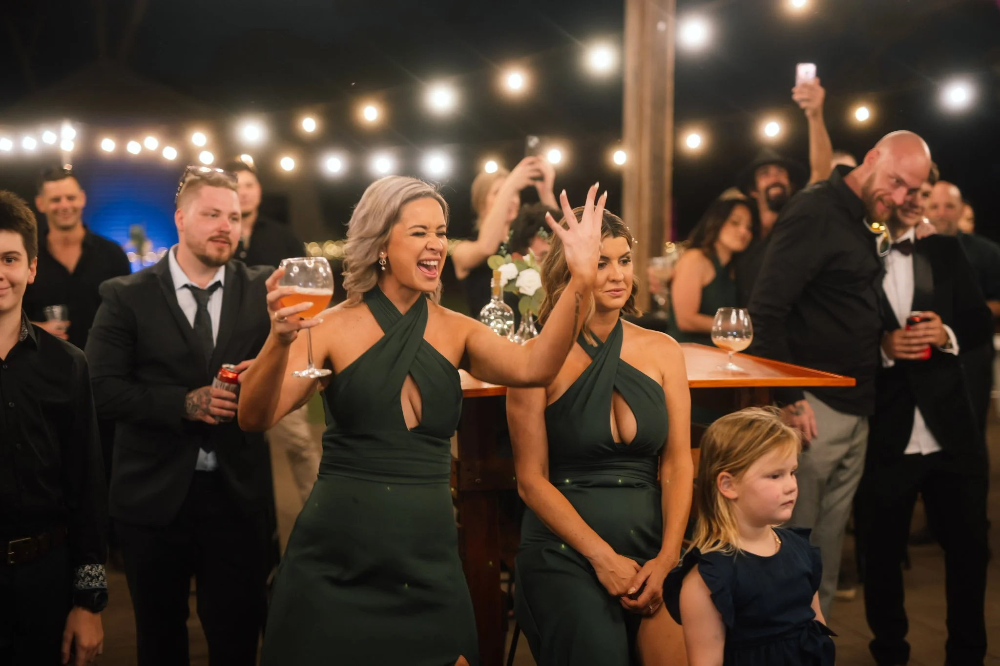 Bridesmaids in elegant emerald green dresses cheering dynamically and raising their drinks under outdoor festoon lights at a Buckland Estate wedding reception.