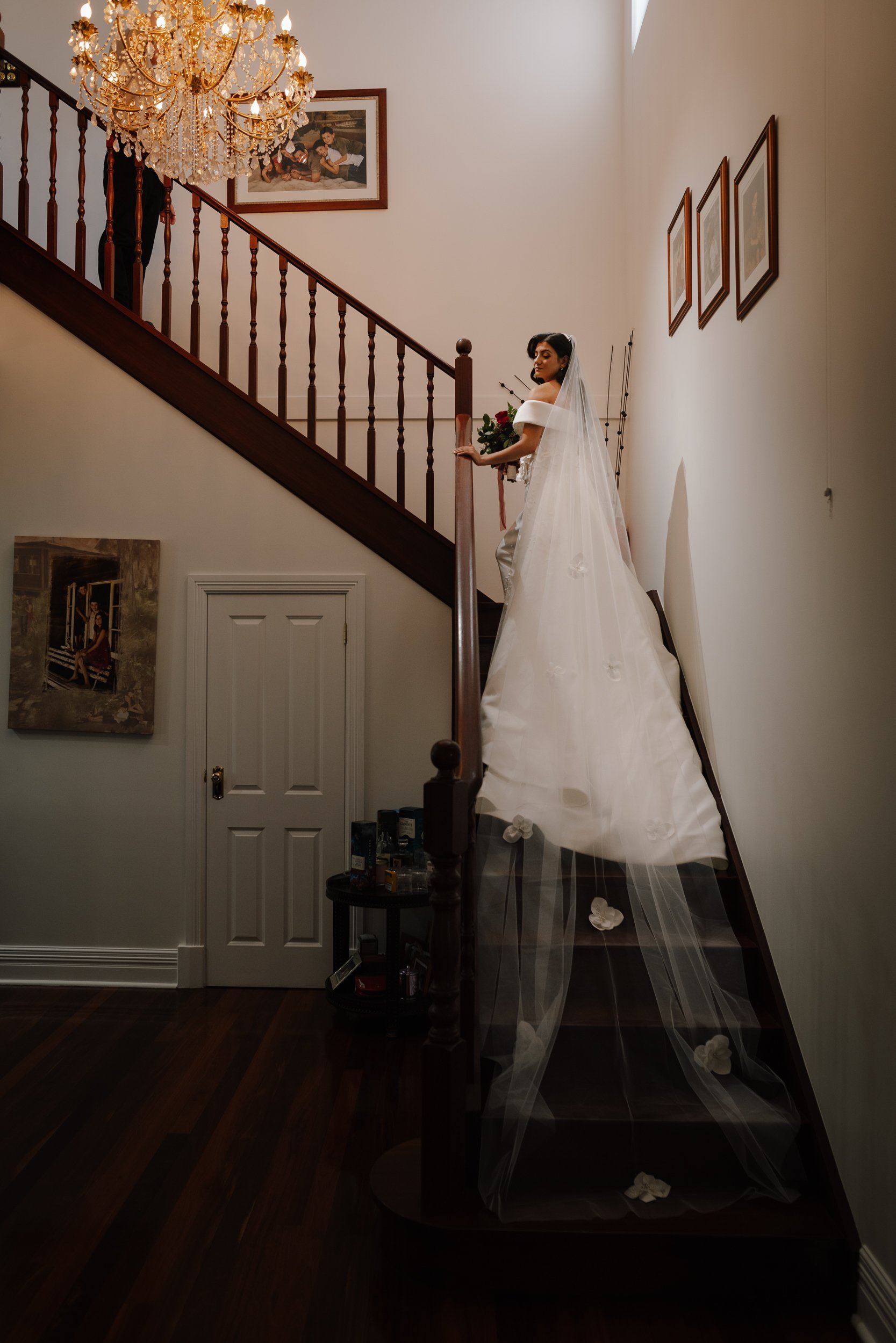 A bride in an off-shoulder gown with a flowing floral-appliqué cathedral veil descends a dark timber staircase