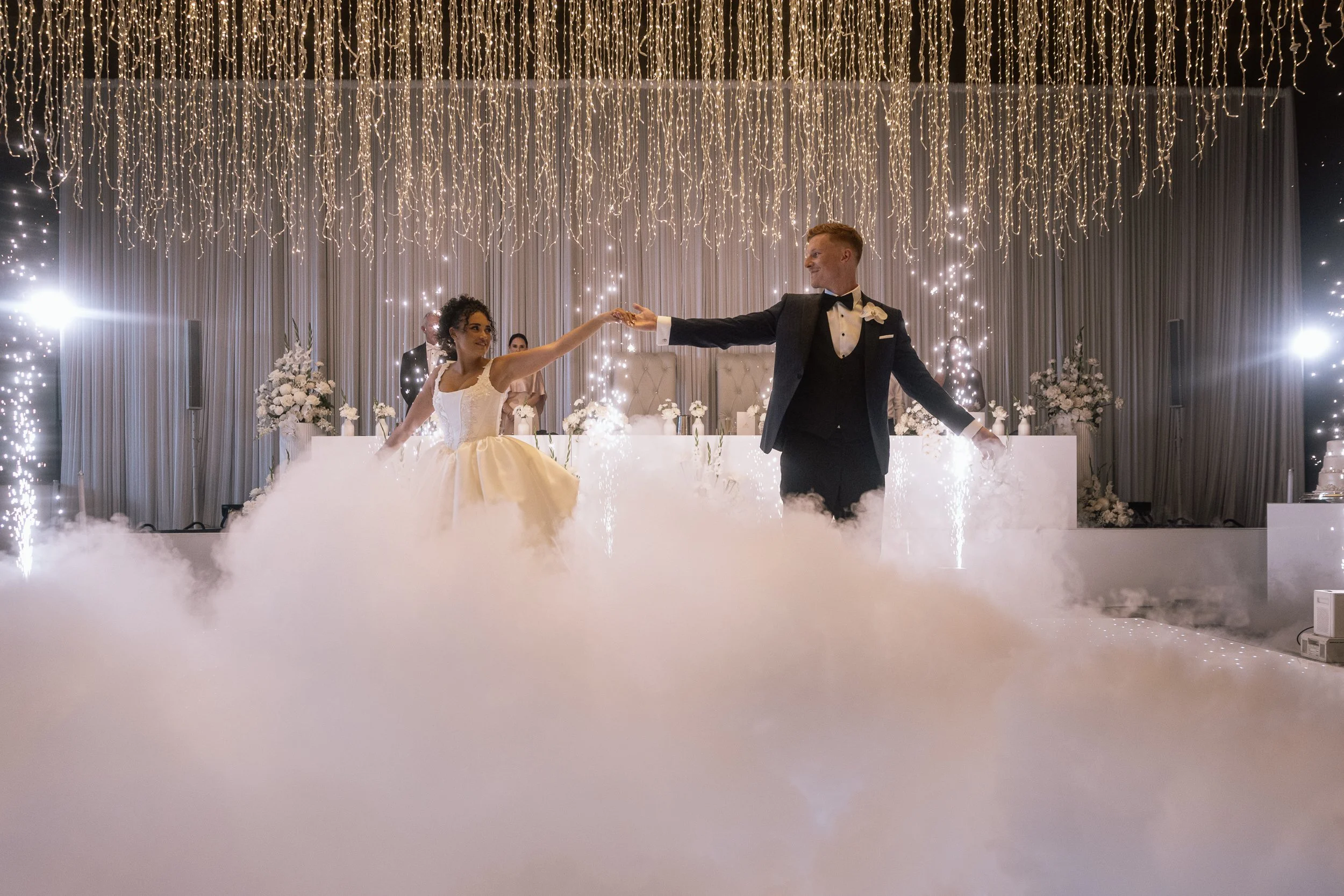 Bride and groom hold hands during their first dance, dry ice fog swirling at their feet beneath a cascading fairy-light