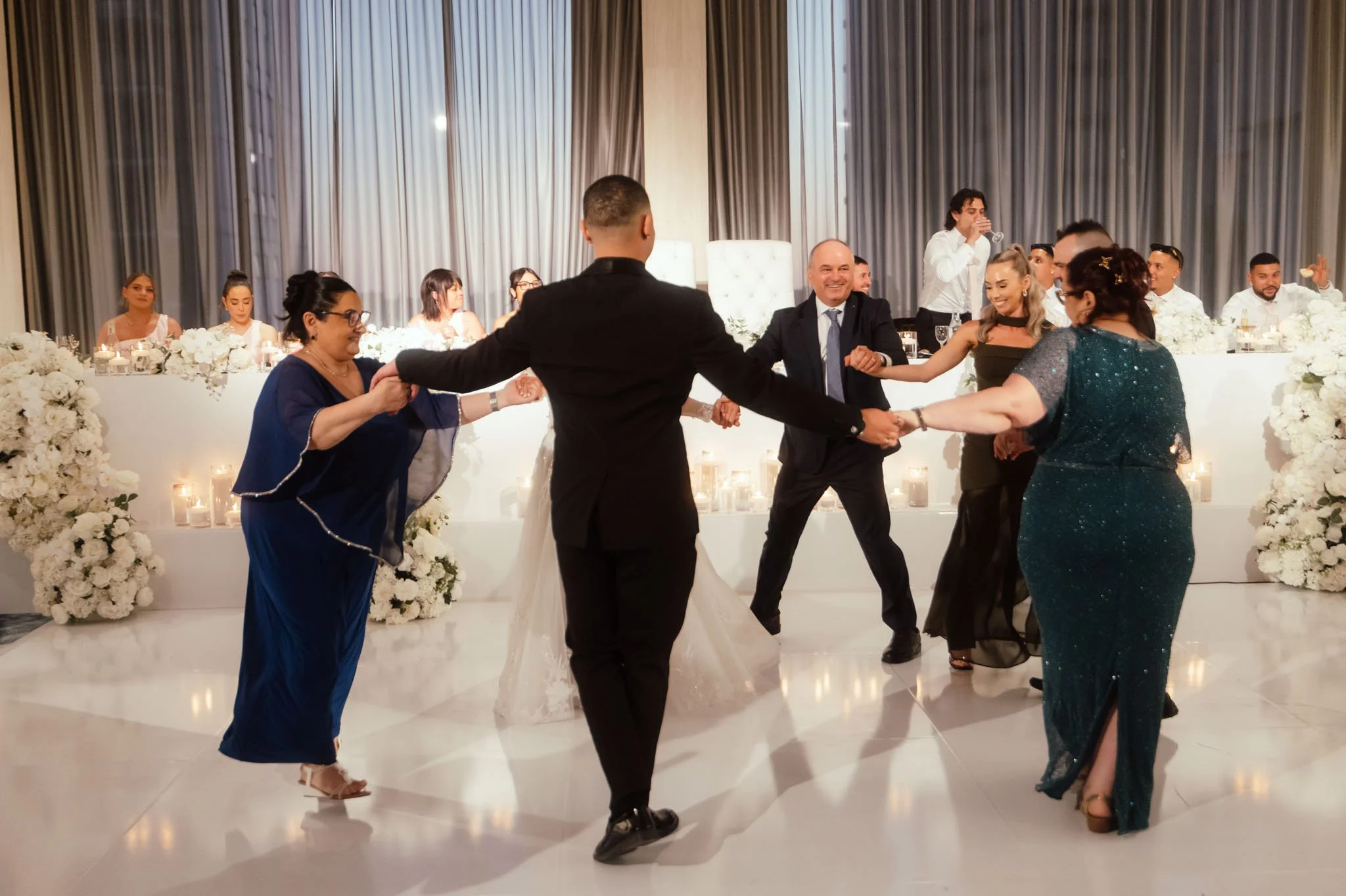 Groom dancing joyfully in a circle with family members on the white dance floor at their Crown Perth wedding reception, with the illuminated white bridal table and white floral arrangements in the background, photographed by James Di Stefano