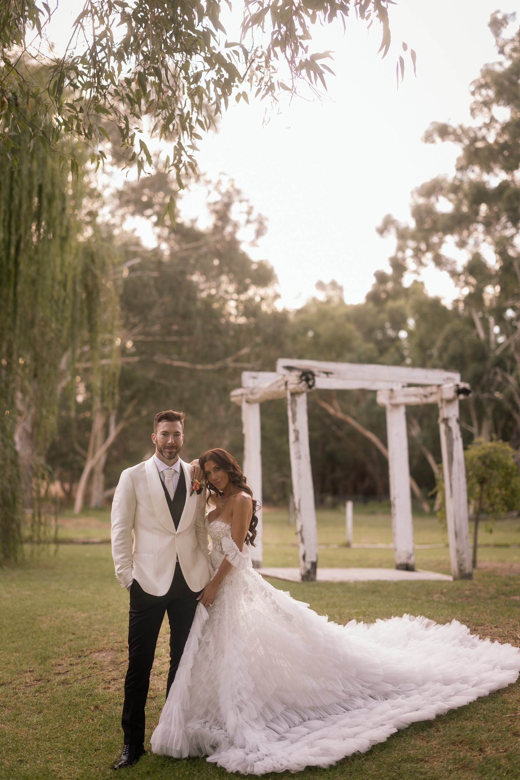 Bride in a voluminous ruffled tulle gown with long train leans beside her groom in a white dinner jacket, a weathered