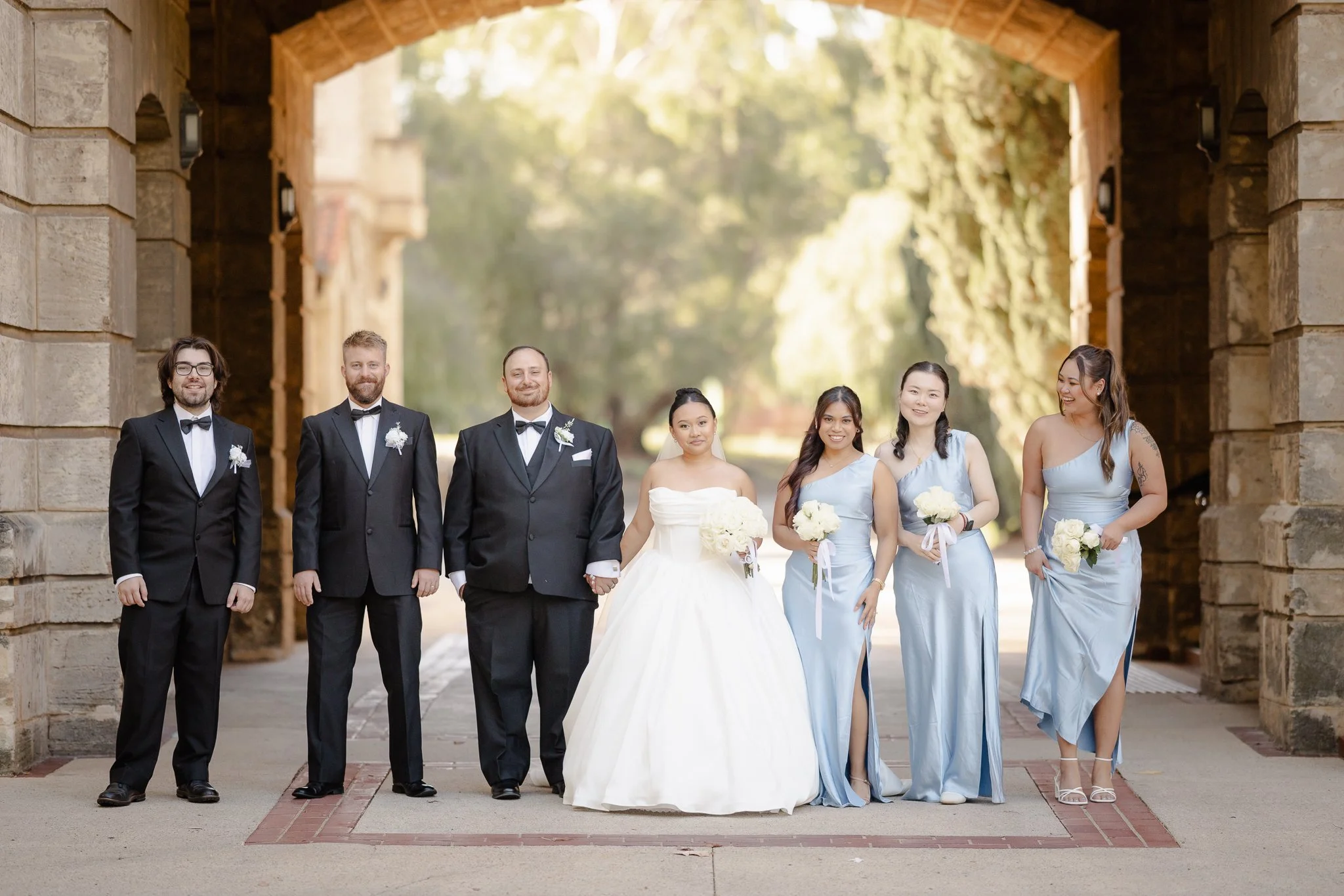 Bridal party in powder-blue gowns and black tuxedos posed beneath a grand sandstone archway, Perth wedding photography.