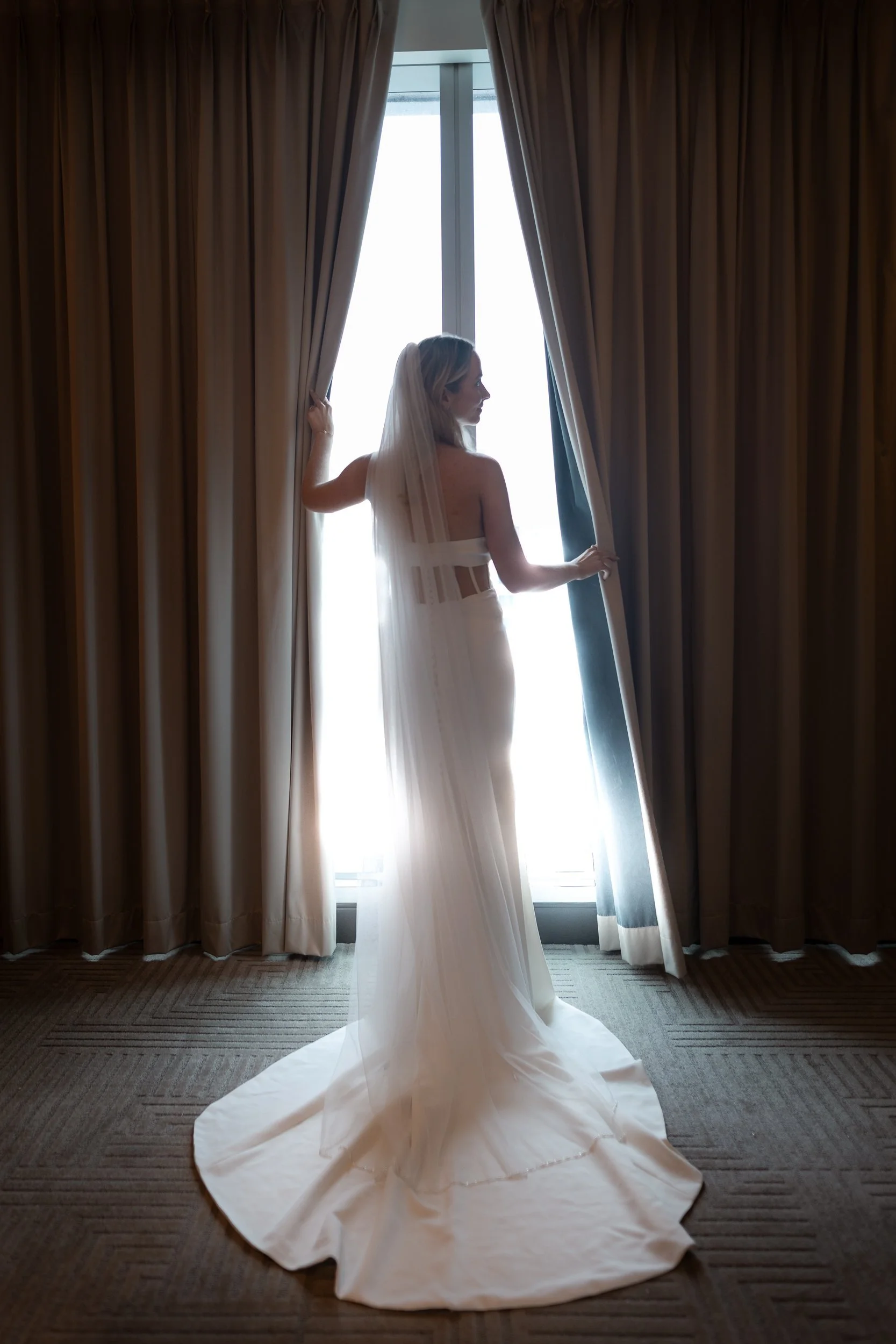 A bride in a sleek fitted wedding gown and cathedral veil parts heavy curtains, bathed in soft window light.