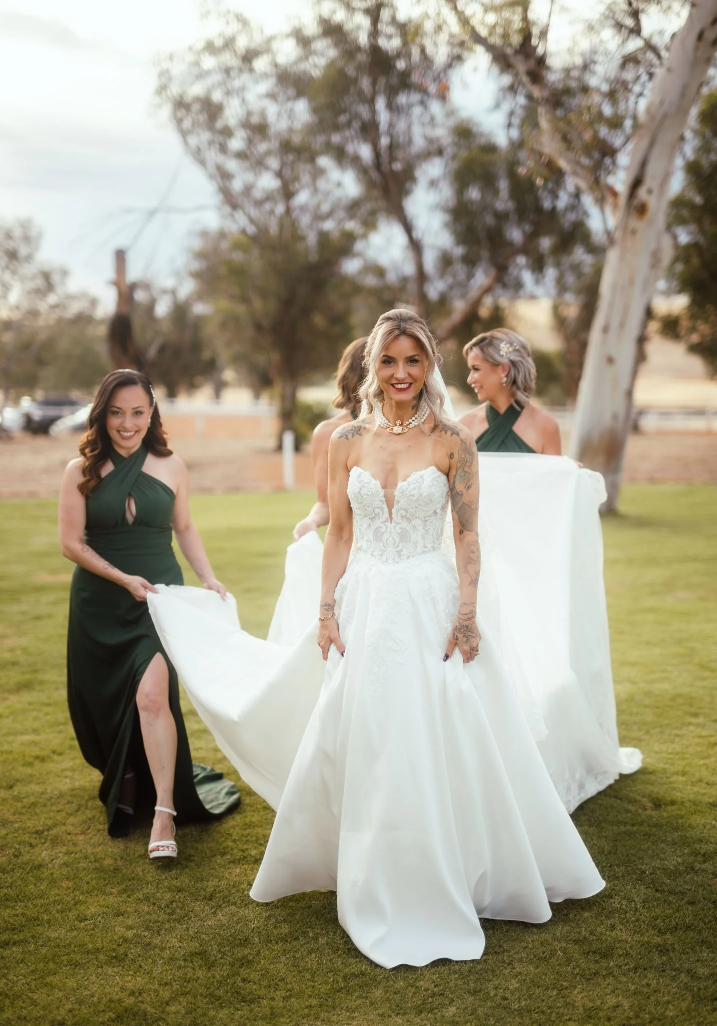 A cheerful, candid photograph of the bride walking across the expansive lawns of Buckland Estate, accompanied by two bridesmaids in long emerald green dresses who are helping her hold her gown