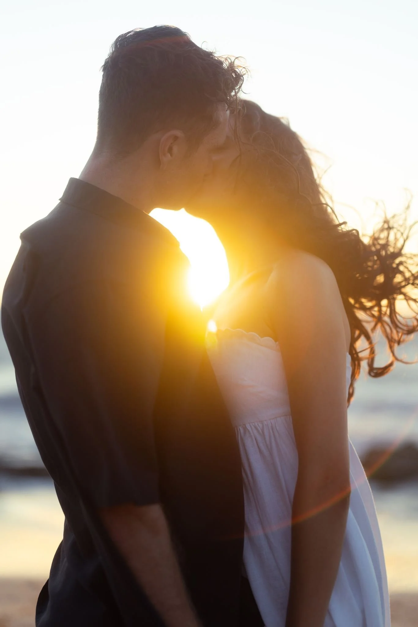A couple share a golden-hour kiss on the beach, the setting sun blazing between them and her hair caught in the coastal breeze