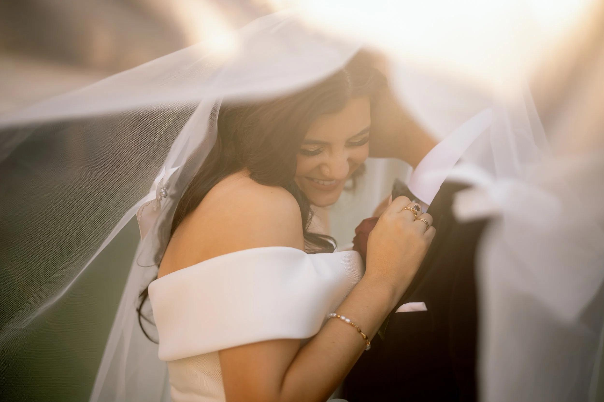 Smiling bride in an off-shoulder gown pulls her groom close beneath a flowing veil, bathed in warm golden-hour light.