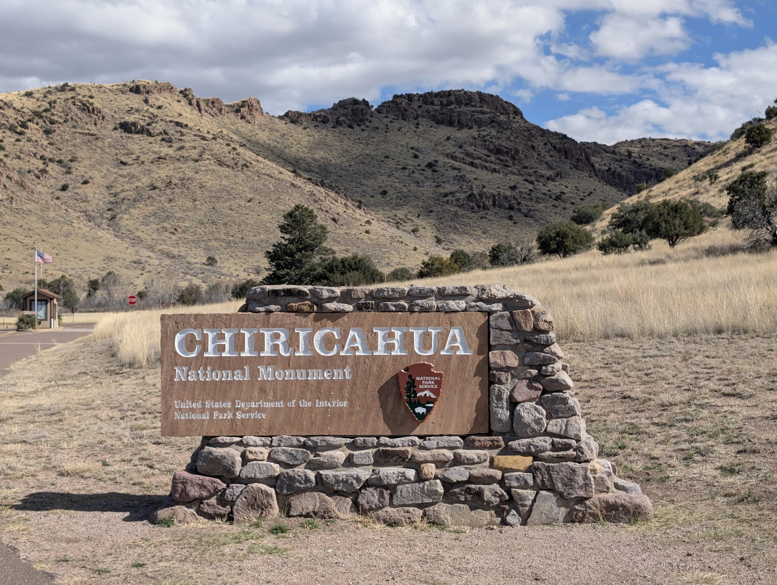 Beautiful wooden sign for Chiricahua Monument in foreground with hills and rock formations in background. Blue skies and white clouds are above.