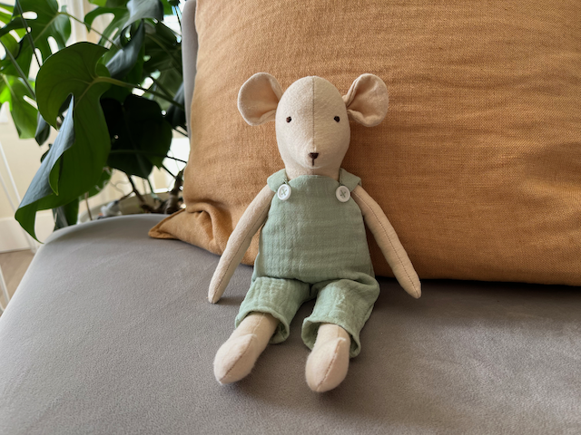 A handmade toy mouse with green overalls sits on a couch.