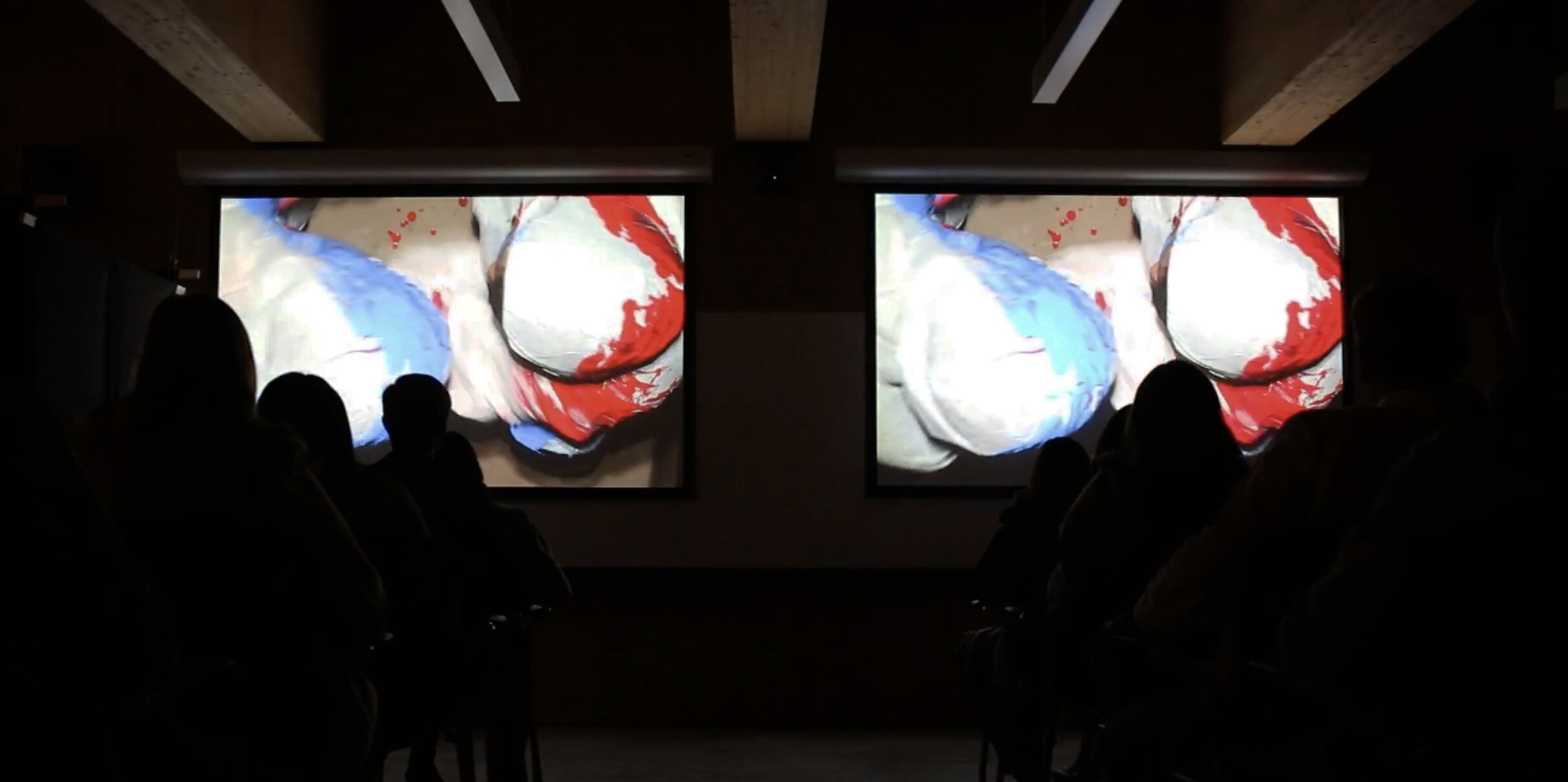  In this image the authors are filmed from above applying the blue and red paint to each other. The intimate shared struggle of the authors/performers is here mediated for the audience through the lens of a camera installed on the ceiling. Photograph