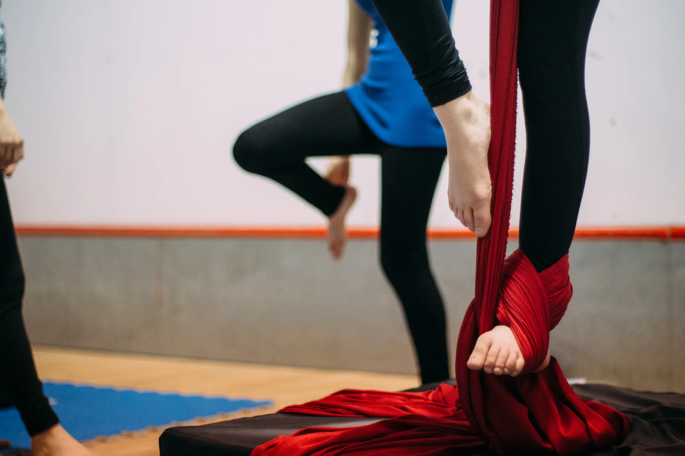 Person practicing aerial silk acrobatics on red fabric, with another person in the background on a yoga mat in a studio.