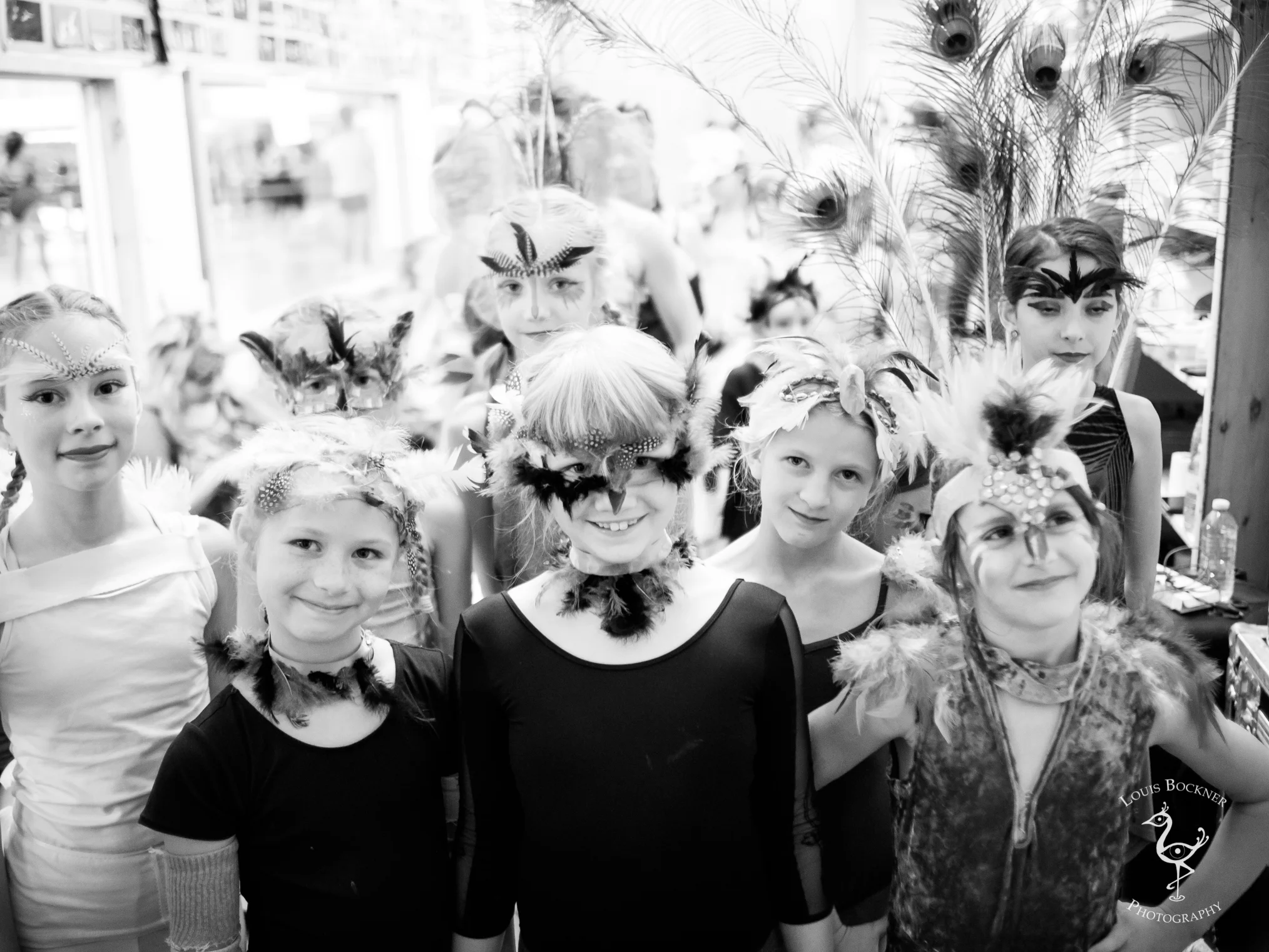 A group of young girls dressed in feathers and face paint, likely for a costume or dance performance, standing indoors with a background of peacock feathers and decorations.