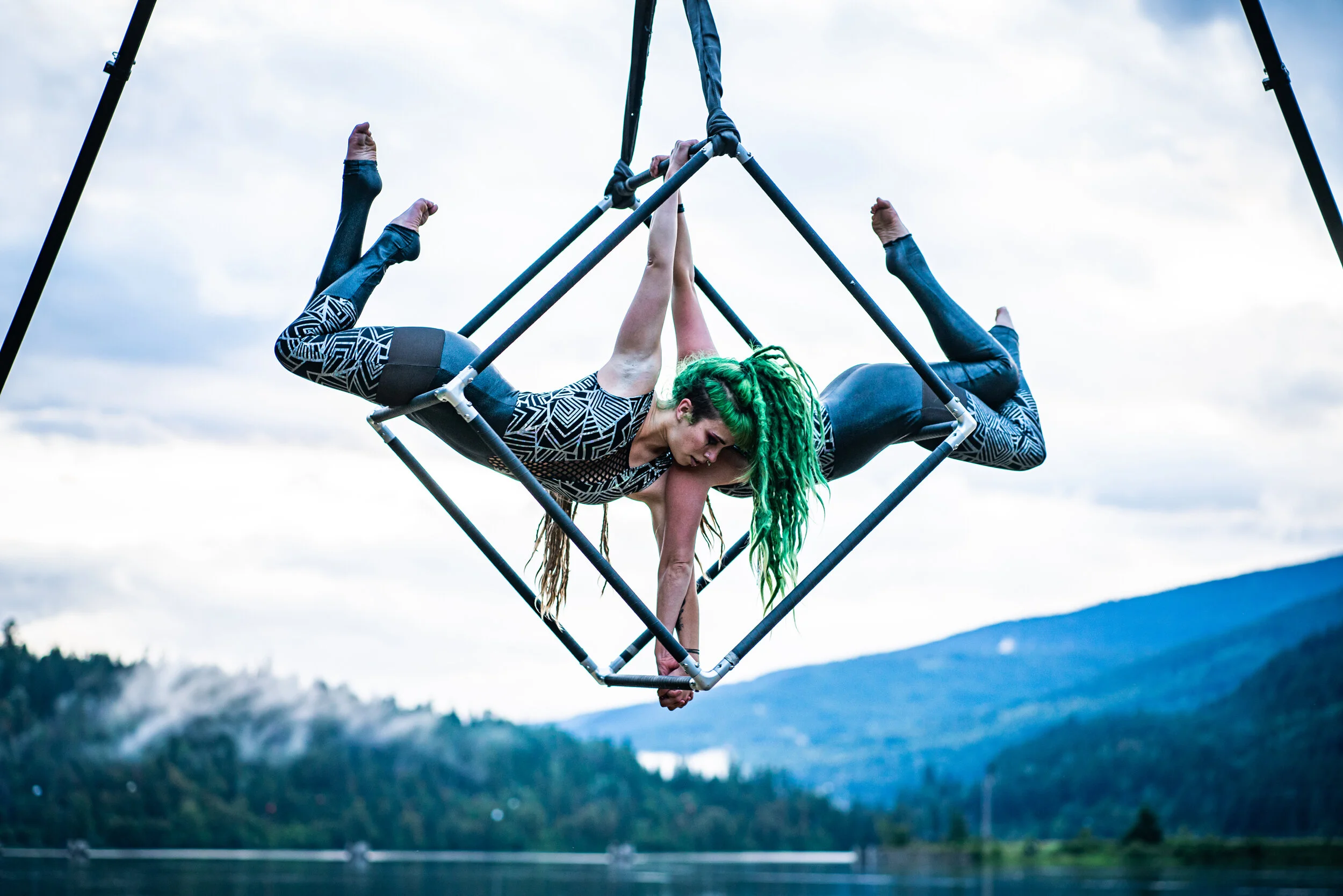A woman with green dreadlocks performing acrobatics on a metal cube structure outdoors with a lake and mountains in the background.