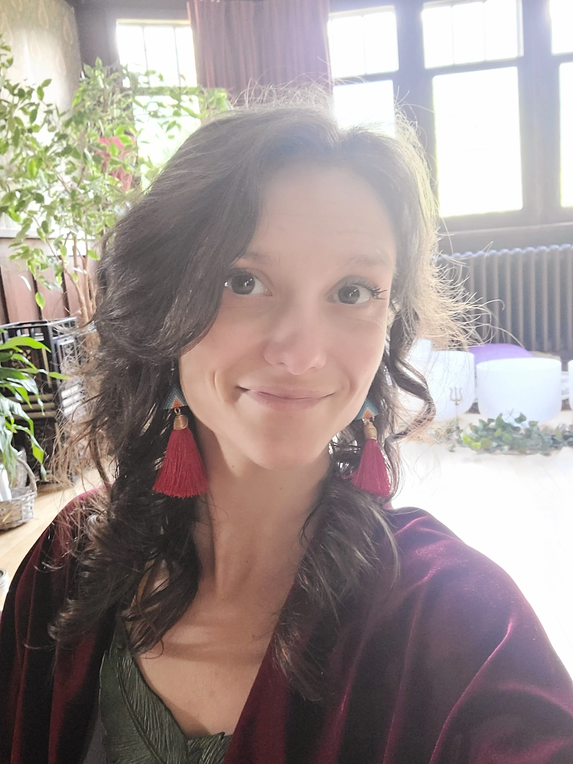A woman taking a selfie indoors, with natural light coming through large windows behind her. She has dark wavy hair, wearing red tassel earrings and a maroon velvet jacket, with a smile.