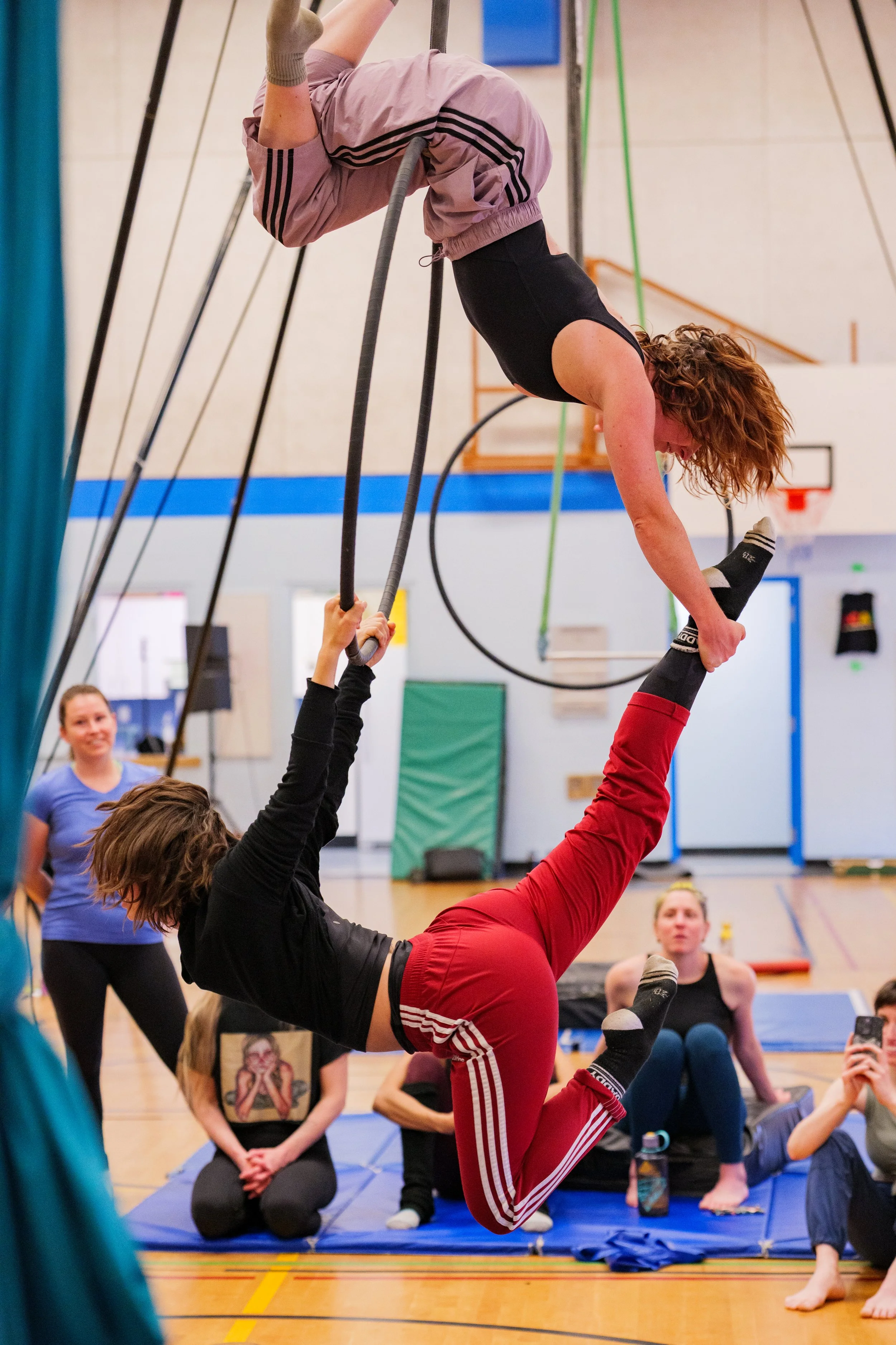 A woman practicing acrobatics upside down on gymnastic rings with the help of a spotter holding her legs, in a gymnasium with spectators sitting on mats and watching.