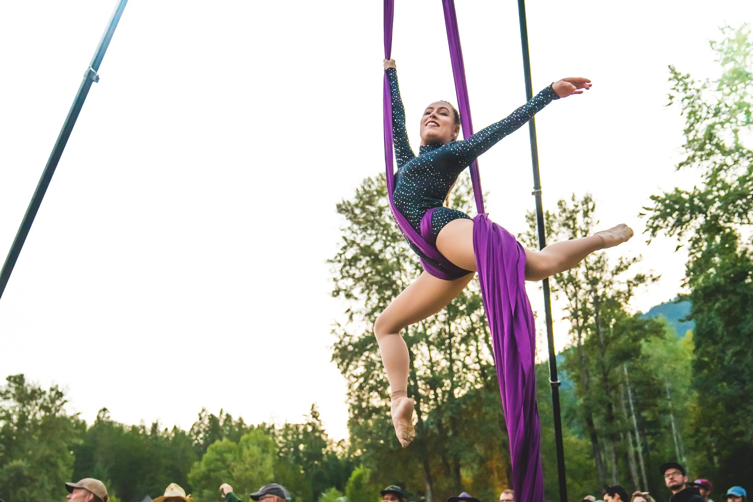 A female acrobat performs aerial silk act outdoors during the day, suspended in the air with purple silks, smiling and extending her arms and legs in a dance pose, with greenery and onlookers in the background.