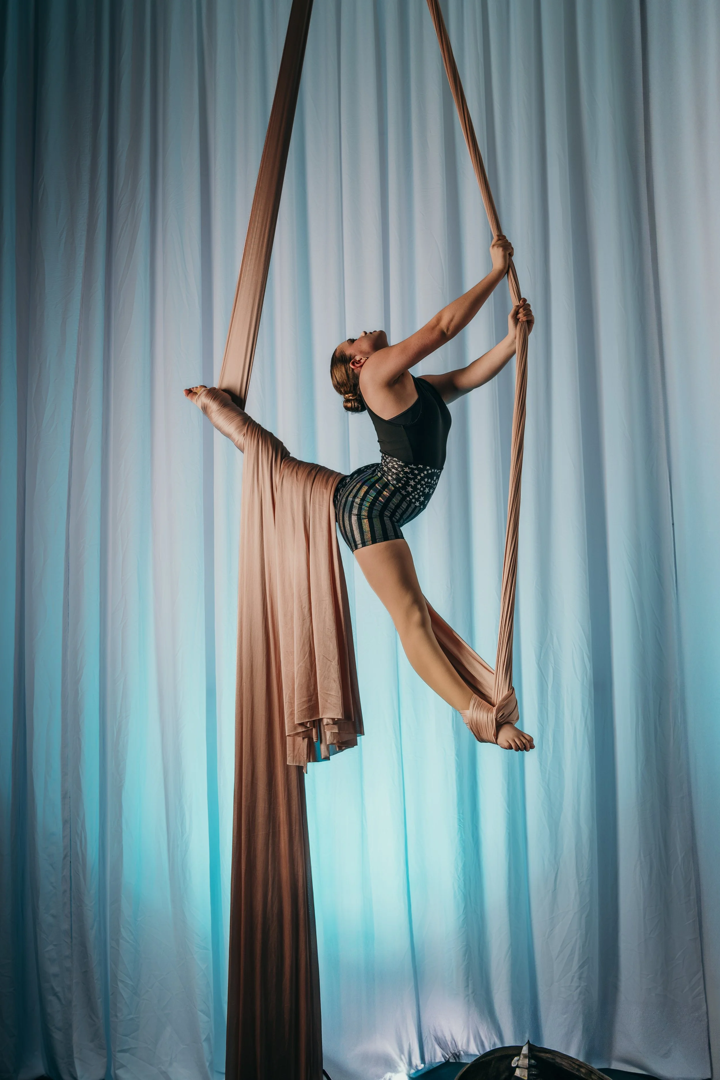 Aerial silk performer mid-performance, hanging from beige silk fabric in front of light blue curtains.