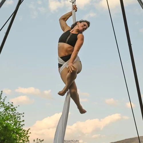A woman performing aerial silk acrobatics outdoors, hanging from a fabric suspended in the air with a cloudy sky background.