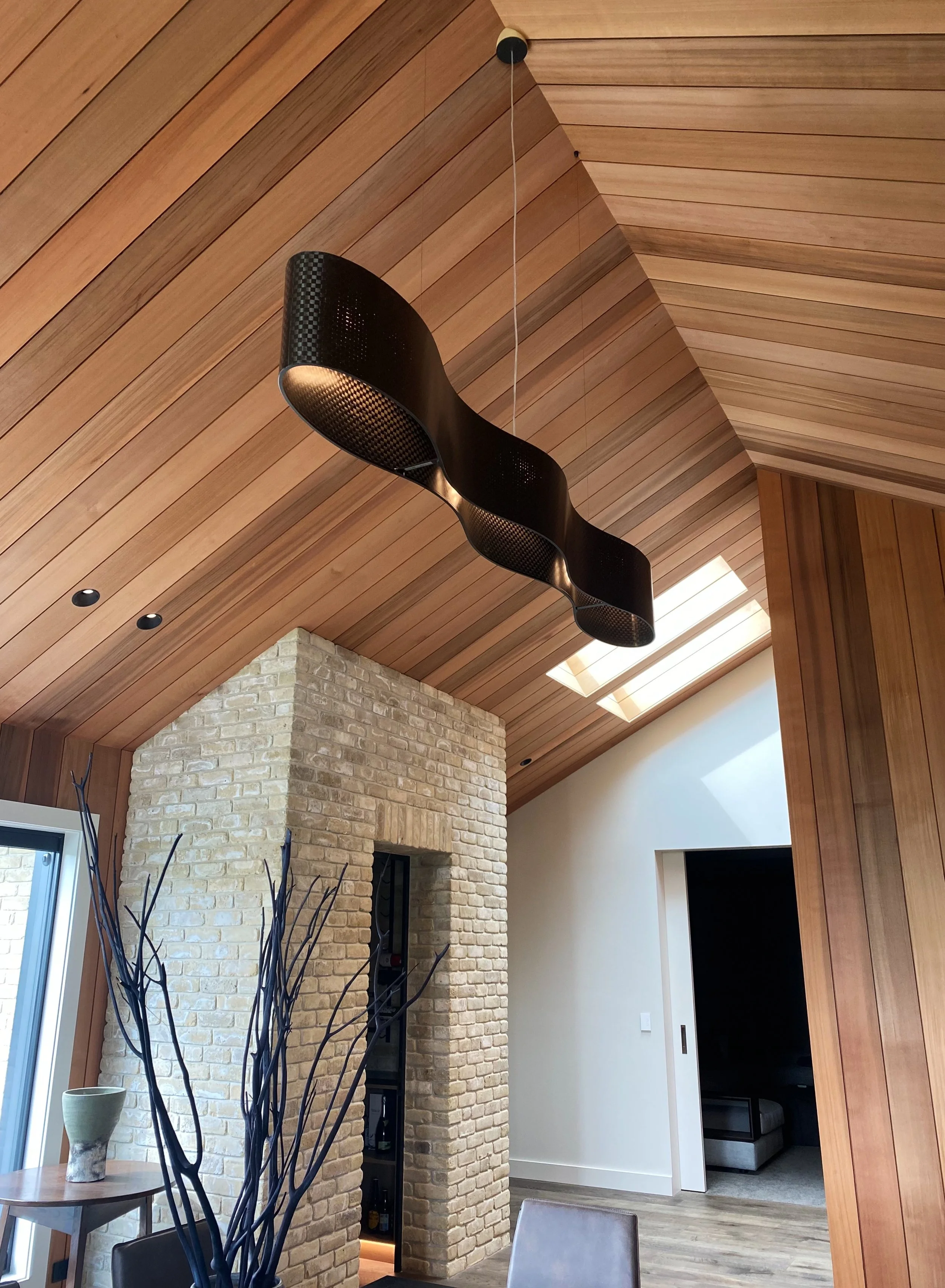 James Russ Infinity Pendant. Interior view of a room with a wooden sloped ceiling, black modern hanging light fixture, brick wall, and a corner with a dining table, chairs, and decorative branches in a vase.
