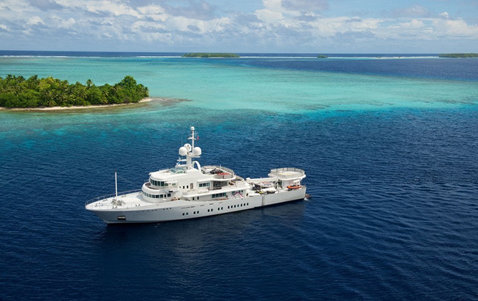 James Russ lighting. A large white yacht anchored near a green tropical island with clear turquoise water, under a partly cloudy sky.