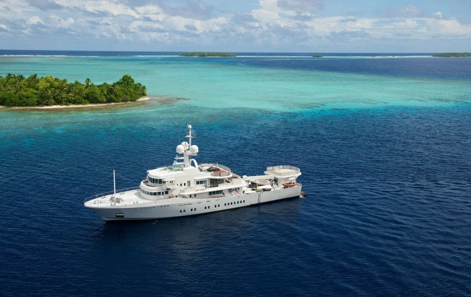 James Russ lighting. A large white yacht anchored near a green tropical island with clear turquoise water, under a partly cloudy sky.