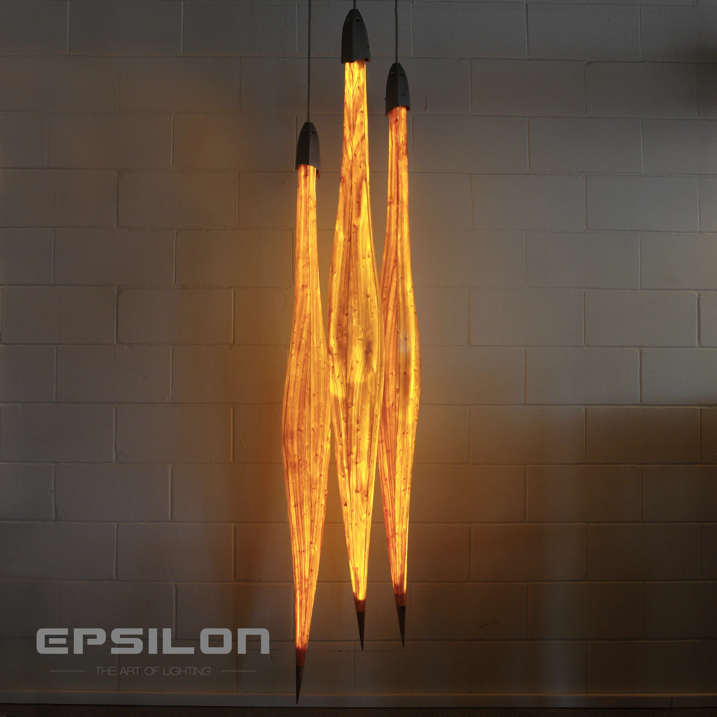 Three elongated, glowing, orange-yellow light sculptures resembling flames or elongated leaves, mounted vertically on a gray brick wall.