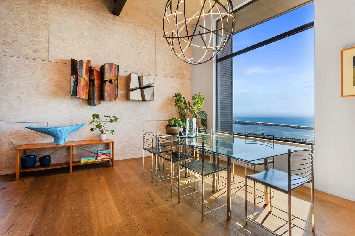 James Russ Atomic Sculpture. Modern dining room with a glass table, six metal chairs, large window with ocean view, decorative wall art, abstract chandelier, and indoor plants.