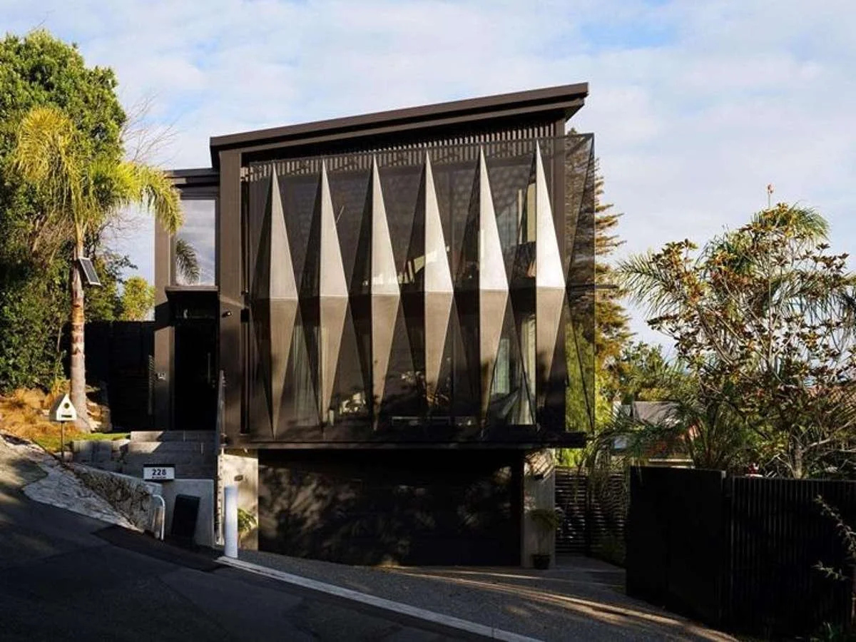 Modern house with angular black exterior and large glass windows, located on a sloped street surrounded by trees.