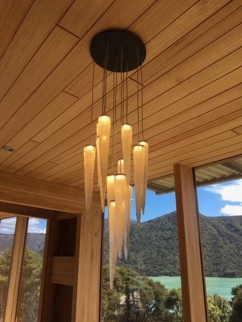 Modern chandelier with elongated light fixtures hanging from a wooden ceiling, overlooking a scenic outdoor view of mountains and a lake.