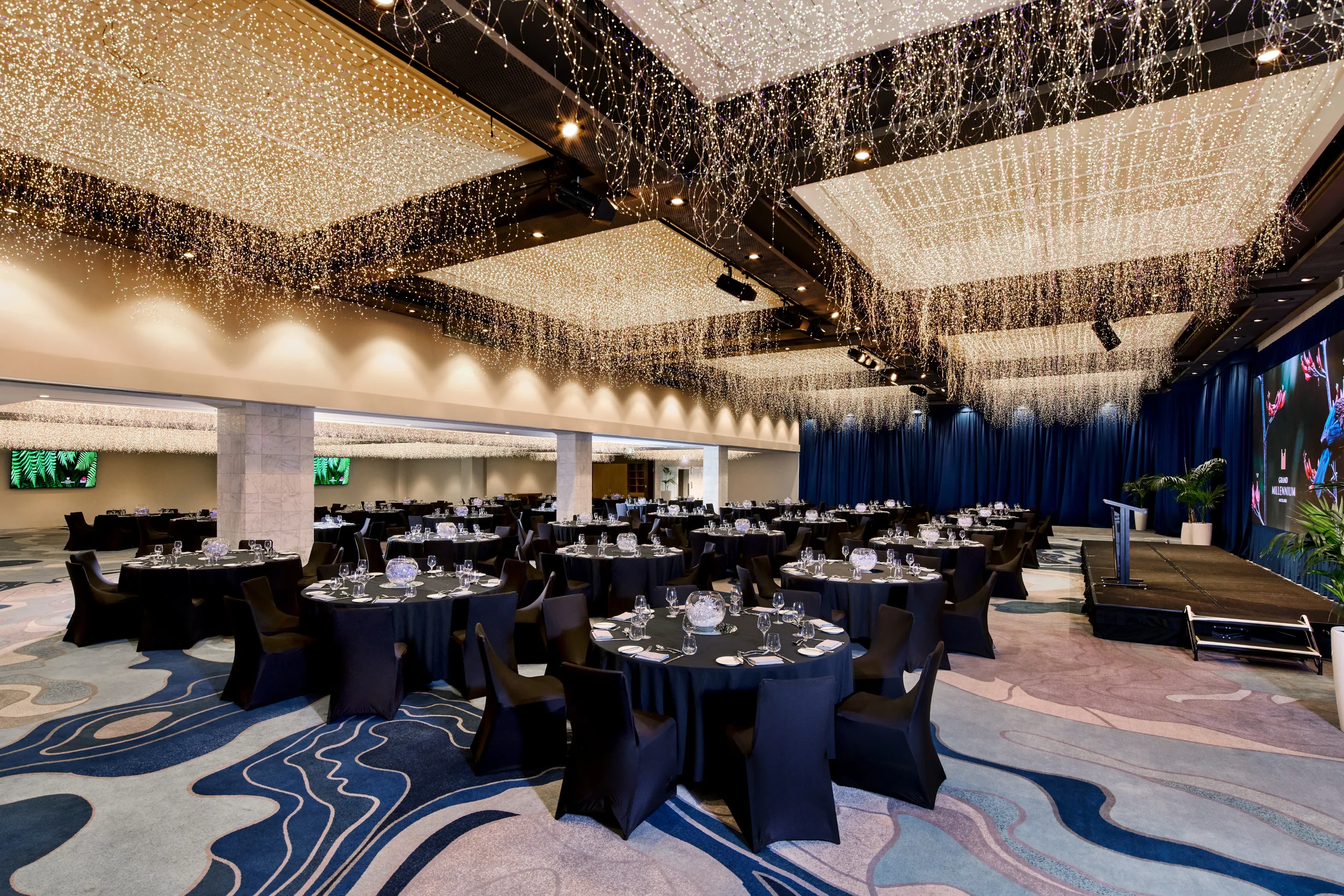 A spacious banquet hall decorated with string lights hanging from the ceiling, black round tables with black chairs, and a stage with a large screen and blue curtains.
