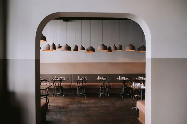 Empty restaurant with hanging james russ pendant lights, chairs, and tables, viewed through an arched doorway.