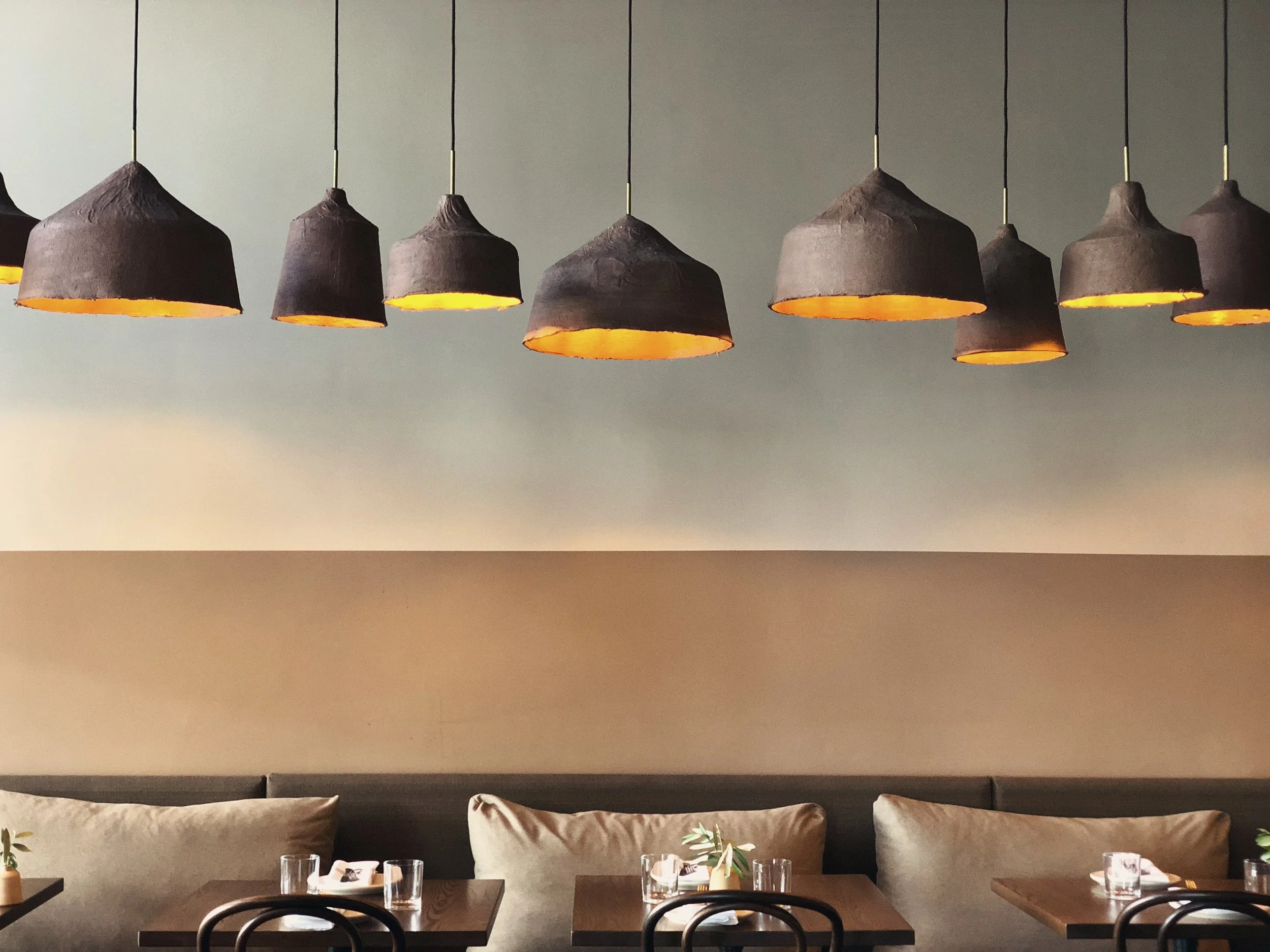 James Russ Andiamo restaurant with a grey wall, modern clay pendant lights with an orange glow, beige cushioned seating, and a dining table with glasses and a small plant.