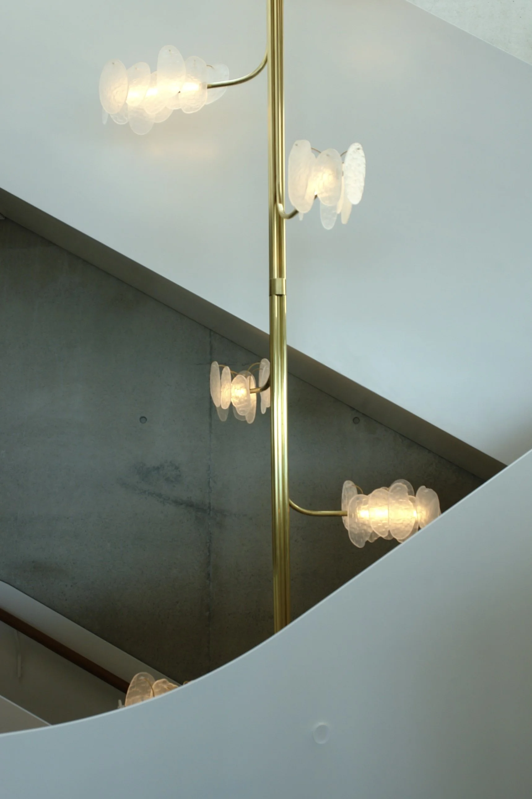 Interior view of a modern staircase with floating glass light fixtures attached to a vertical brass-colored pole, viewed from below, featuring concrete walls and a ceiling with geometric shapes.