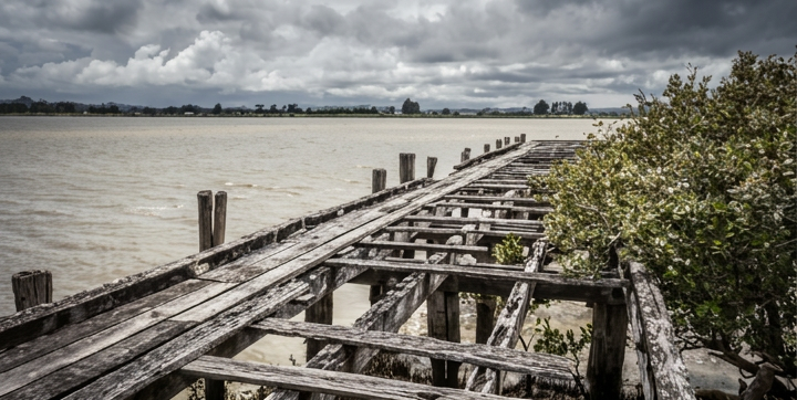 James Russ Portland wharf Old, weathered wooden pier extending over a body of water, with a cloudy sky overhead and bushes on the right side.