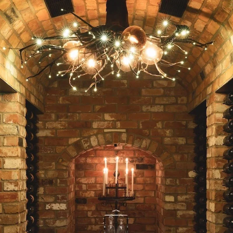 A brick wine cellar with a chandelier made of branches and bulbs on the ceiling, and a wall-mounted candelabra with three lit candles on the back wall. Wine bottles are stored on shelves along the sides.