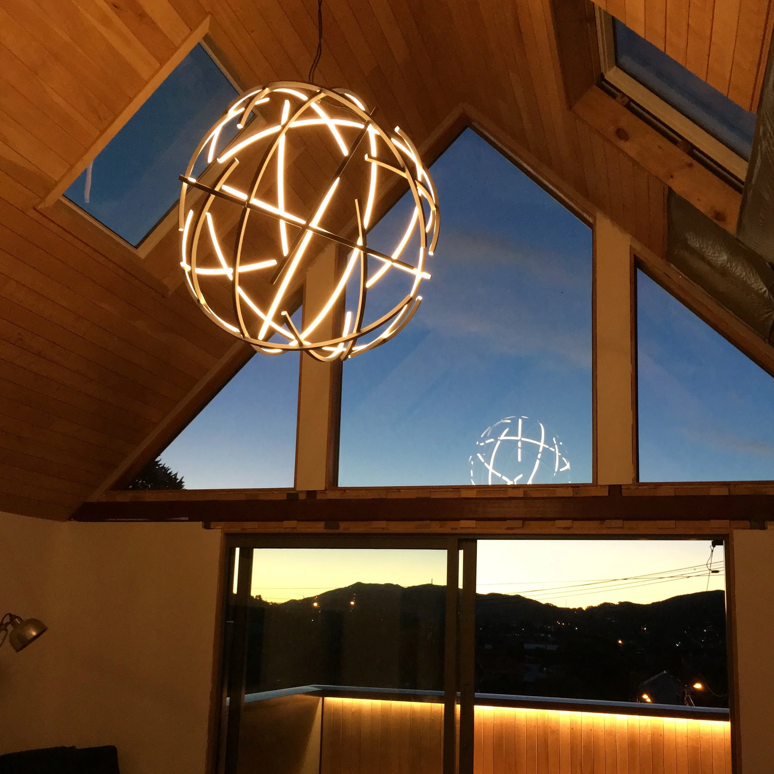 Interior view taken during dusk showing a spherical chandelier hanging from wooden ceiling beams, with three rectangular windows exposing a landscape of hills and evening sky.