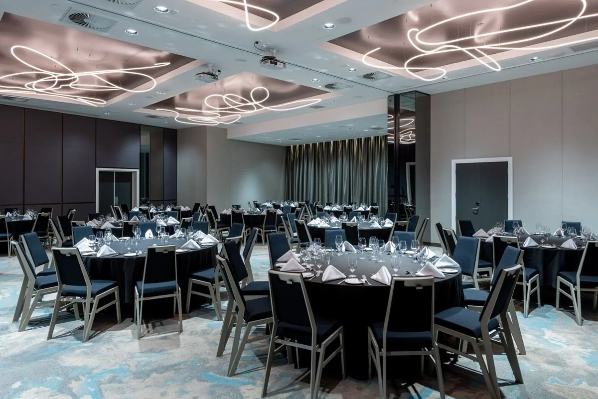 A banquet hall with round tables covered in black tablecloths, set with glassware, napkins, and silverware, illuminated by modern ceiling lights, with dark chairs arranged around each table.