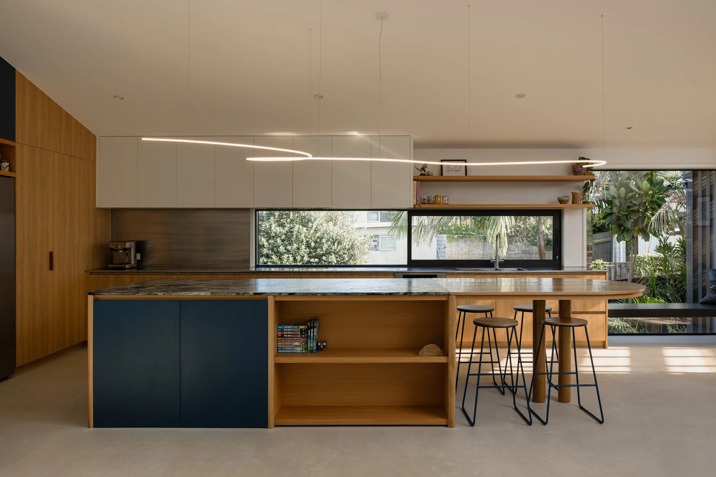 Modern kitchen with a wooden island, navy blue cabinets, and a marble countertop. Open shelving with plants, books, and decorative items. Large window with greenery outside, and contemporary LED pendant lighting.