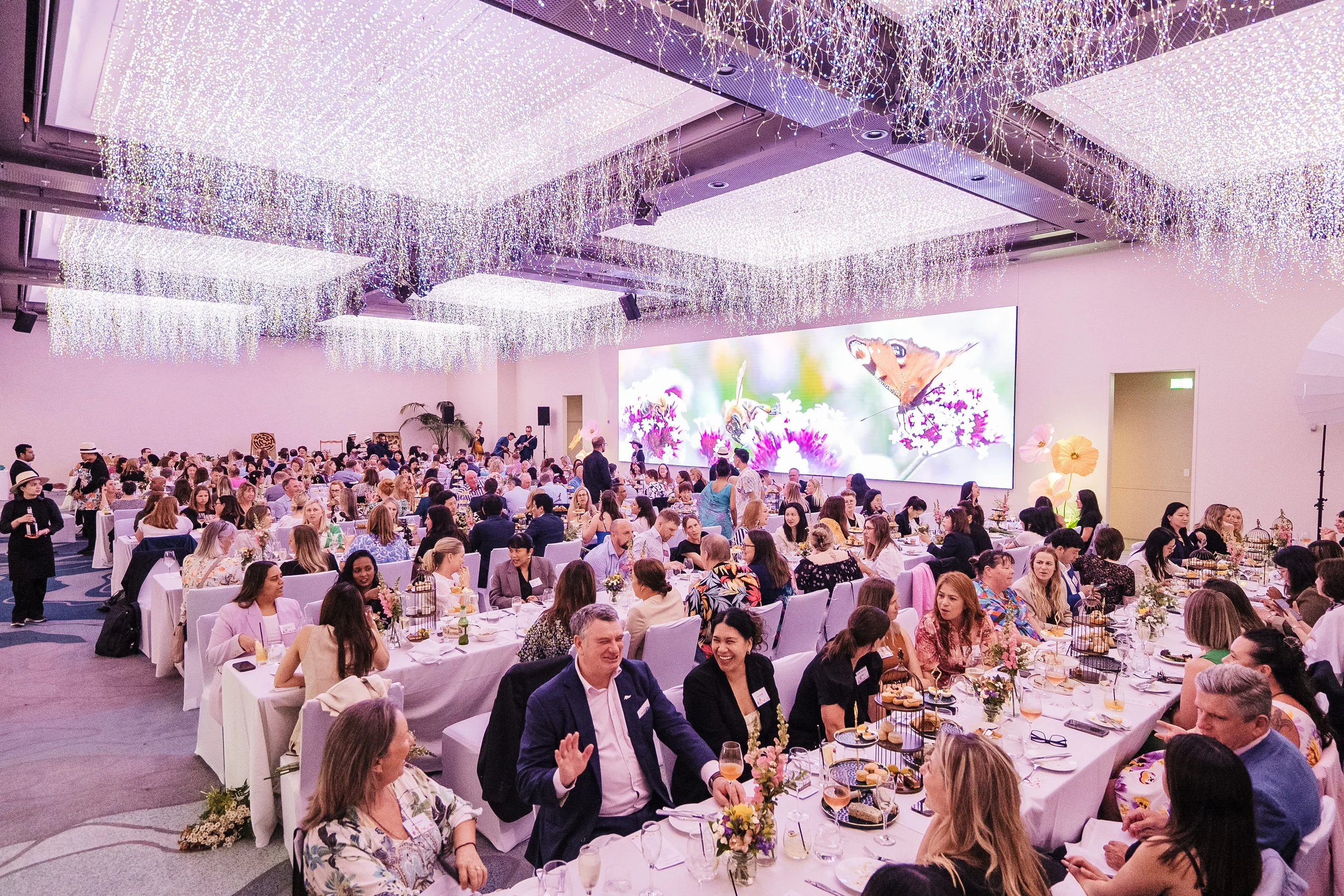 A large banquet hall filled with people dining at decorated tables for a formal event, with a large digital screen displaying a butterfly and floral image on the grey wall in the background, and curtains of cascading LED lights on the ceiling.