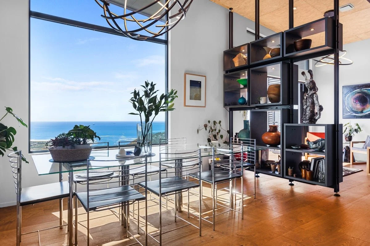 James Russ Atomic Sculpture. Modern dining room with a glass table and six metallic chairs, large window with ocean view, wooden floor, contemporary shelving with sculptures and decorative items, potted plants, and abstract art.