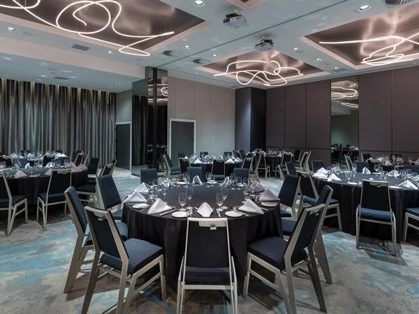 James Russ Sheraton event hall set for a formal event, featuring black tablecloths, white napkins, wine glasses, and silverware. Modern ceiling lights with abstract designs and large mirrors reflect the room's decor.