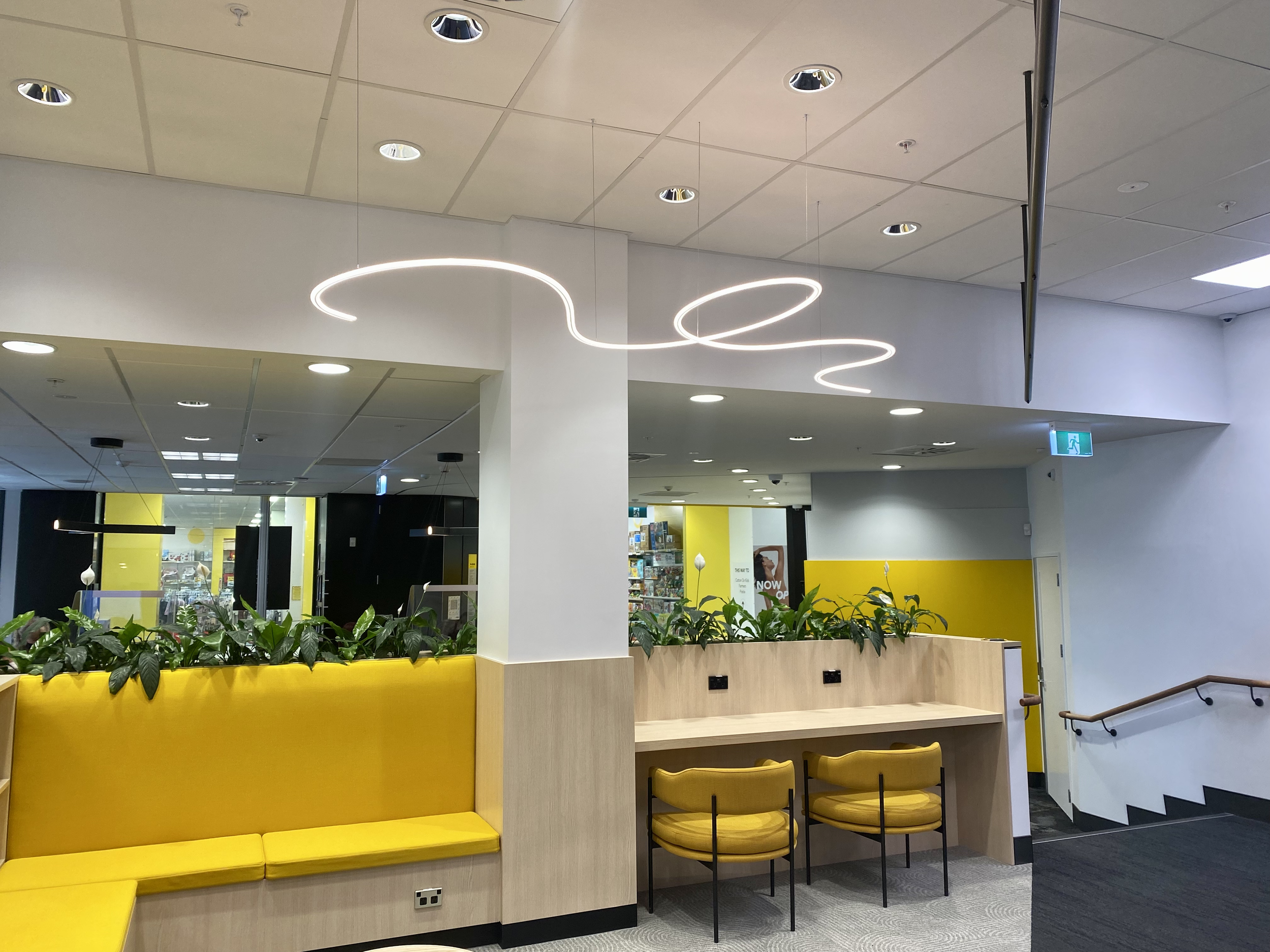 James Russ Sensory Sculpture. Modern interior space with yellow seating area, green plants, high ceiling with circular light fixtures, and a hanging abstract LED light fixture.